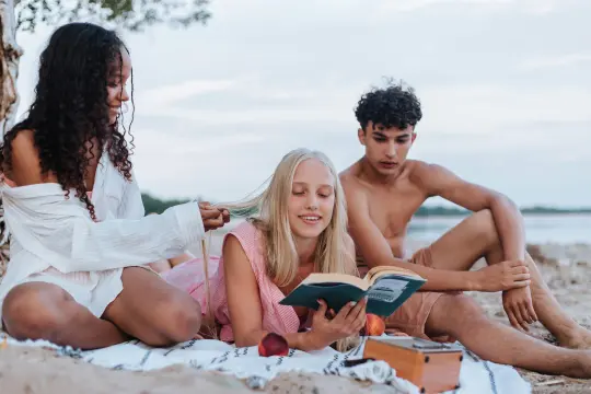 Tre unge mennesker sitter/ligger på en strand. Personene i midten holder en oppslått bok. 
